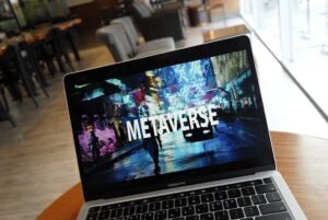 A laptop on a wooden table displaying the word "metaverse" on its screen, symbolizing the concept of virtual reality worlds alive with lifestyle, with a blurred background of a cafe setting.